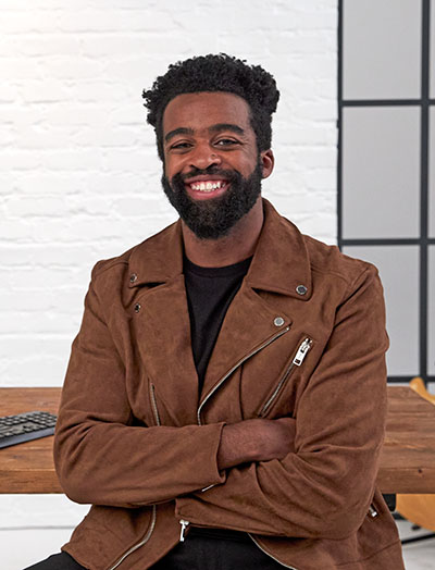 Web design professional sitting at his desk with his arms are crossed and there's a guitar and a monitor in the background. The monitor is displaying the Partner Program website.